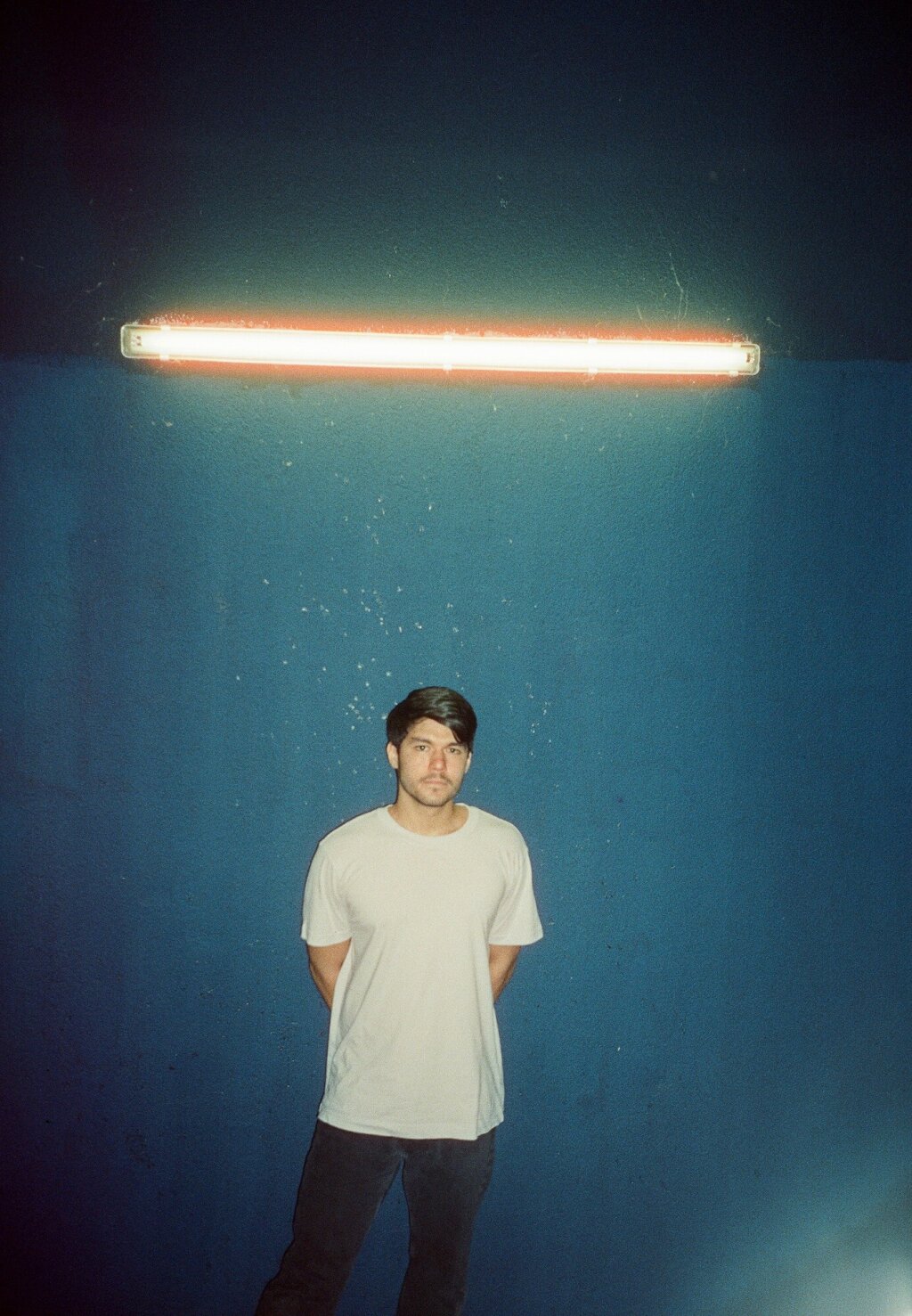 man in a white t-shirt standing in front of a blue wall, illuminated by a working light.