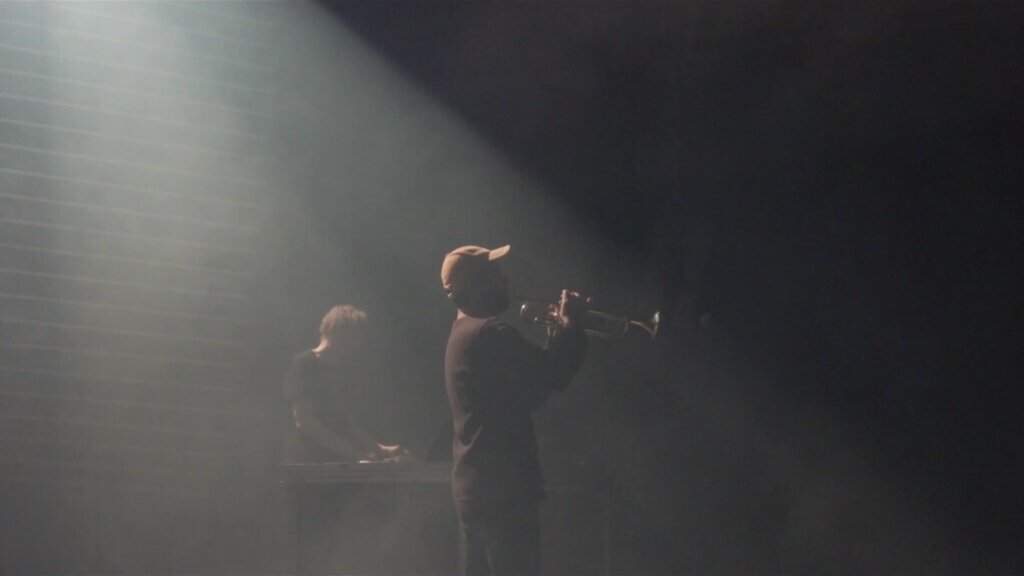 An electronic musician and a trumpeter standing on a foggy stage.