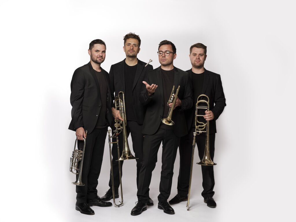 Four wind players in black suits standing on a white backdrop.