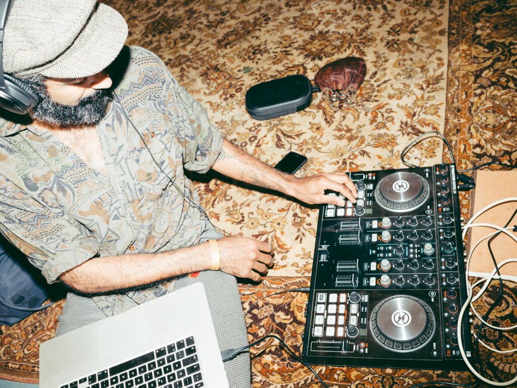 A man sits on a carpet DJing with a controller and laptop.