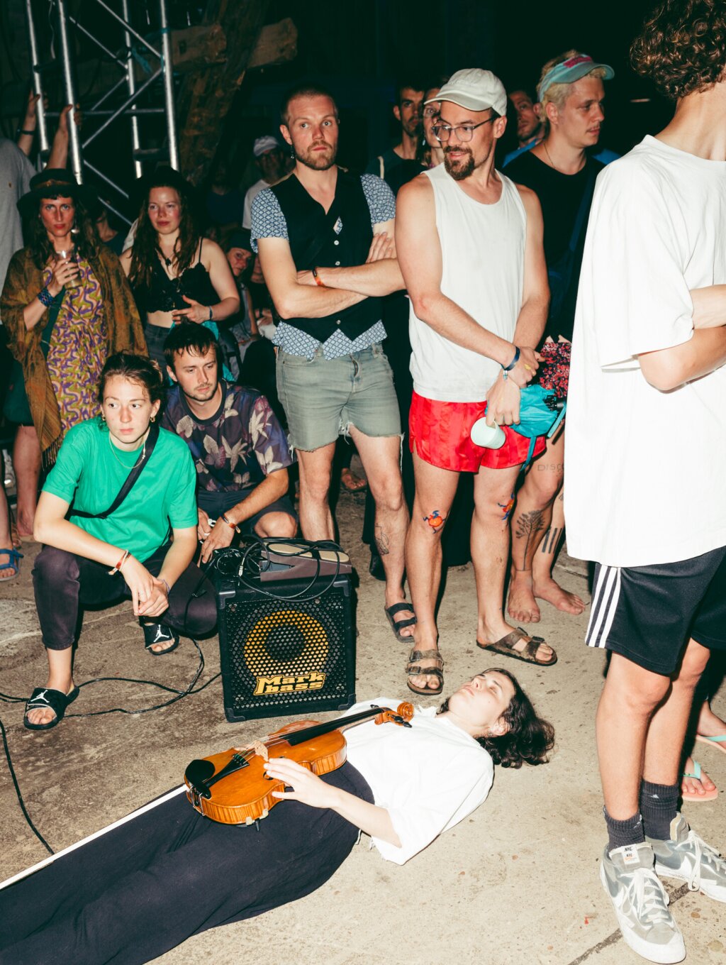 A woman with a violin lies on the floor with the audience in summer clothing standing around her.